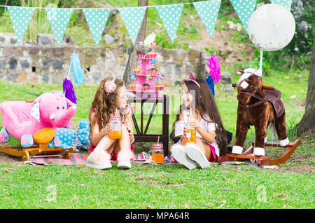 Zwei kleine Mädchen trinken Saft in einer cristal Kolben an draußen im Garten sitzen auf sonnigen Sommertag tragen Pastellfarben Kleidung Stockfoto