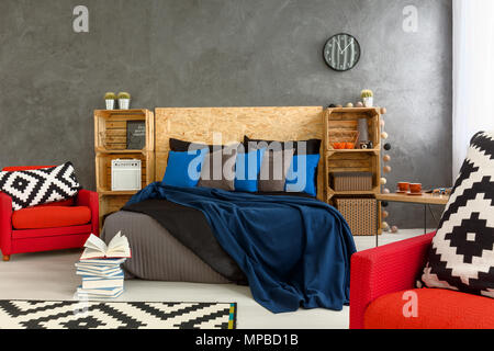 Stilvolle Zimmer mit grauen Wänden und roten Sessel mit wenig Verwirrung auf dem Bett. Holz- Kopfteil des Bettes und Regale aus Holz Boxen Stockfoto