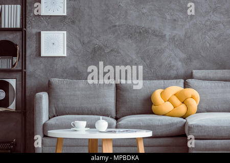 Weiße Tasse auf hölzernen Couchtisch im Wohnzimmer mit gelben Kissen auf graue Sofa gegen die Betonwand Stockfoto