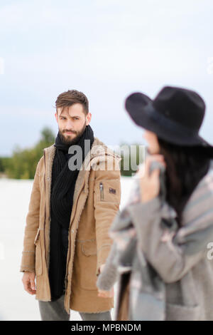 Kaukasische Frau in Hut und Mann mit Bart tragen Mäntel und Schals im Winter schnee Hintergrund stehen. Stockfoto
