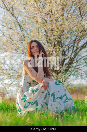 Schlanke sportliche braunhaarige Frau mit blauen Augen in einem blühenden Garten sitzt im Gras in der Sonne Stockfoto