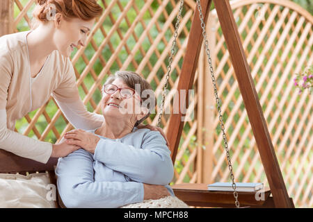 Eine ältere weibliche Rentner mit Behinderungen sitzen auf einer Terrasse Schaukel während der Rehabilitation Camp. Professionelle Hausmeister stand neben der Frau. Stockfoto