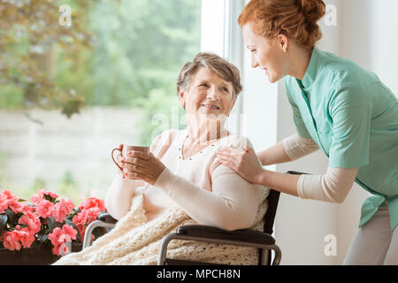 Eine professionelle Hausmeister in Uniform, eine Geriatrische Patientin im Rollstuhl. Senior Holding eine Tasse und Sitzen durch ein großes Fenster in einem REHABI Stockfoto