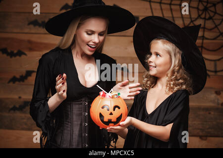 Halloween Konzept - schönen weißen Mutter und ihre Tochter in Hexe Kostüme Halloween Feiern mit sharing Halloween Süßigkeiten und süße über Fledermäuse und Spinnennetz auf Holz- studio Hintergrund. Stockfoto