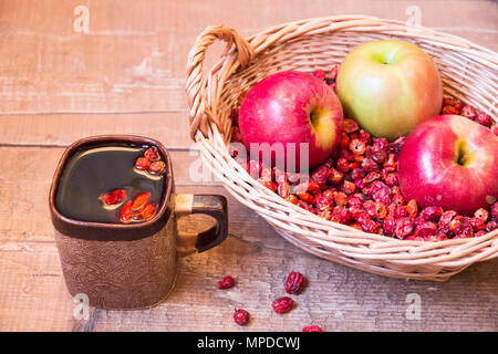 Tasse schwarzen Tee Hagebutten neben Weidenkorb gefüllt mit getrockneten Wild Rose und Äpfel auf Holz- Oberfläche Stockfoto