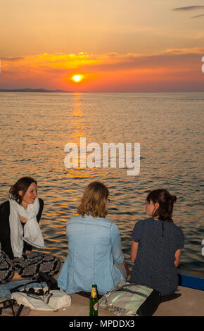 Drei junge Frauen sitzen, trinken und reden am Abend genießen Sie ein herrliches orange Sonnenuntergang vom Wasser in den Hafen von Zadar Kroatien Stockfoto