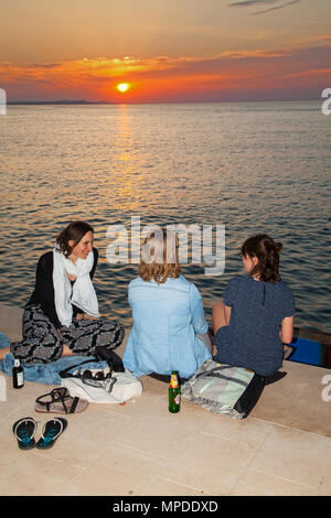 Drei junge Frauen sitzen, trinken und reden am Abend genießen Sie ein herrliches orange Sonnenuntergang vom Wasser in den Hafen von Zadar Kroatien Stockfoto