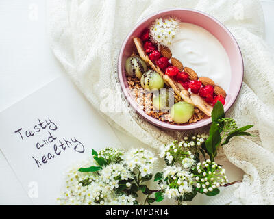 Gesundes Essen. Berry Smoothie, Müsli mit Nüssen, trockene Beeren, Banane und Kiwi in einem rosa Platte, Zweig der wunderschöne, blühende Blumen, noteboo Stockfoto