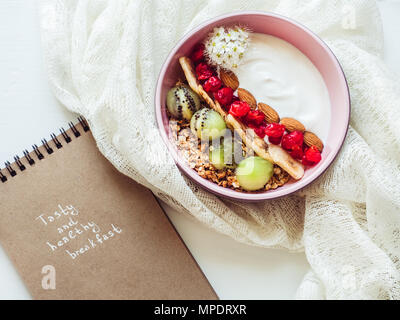 Gesundes Essen. Berry Smoothie, Müsli mit Nüssen, trockene Beeren, Banane und Kiwi in einem rosa Platte, Zweig der wunderschöne, blühende Blumen, noteboo Stockfoto