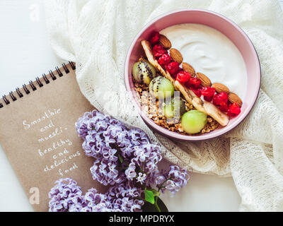 Konzept der gesunden Ernährung. Berry Smoothie, Müsli mit Nüssen, trockene Beeren, Banane und Kiwi in einem rosa Platte, schöne, blühende Blumen, notebo Stockfoto