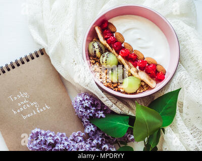 Konzept der gesunden Ernährung. Berry Smoothie, Müsli mit Nüssen, trockene Beeren, Banane und Kiwi in einem rosa Platte, schöne, blühende Blumen, notebo Stockfoto
