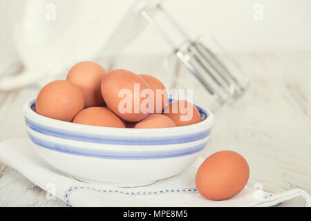 Käfig frei, braune Eier in eine weisse und eine blaue Schale am Küchentisch. Eine Hand Mixer im Hintergrund. Stockfoto