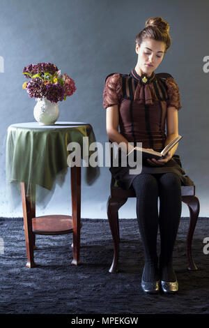 Junge Frau in Vintage-Mode Bluse sitzt neben dem Tisch mit Blumen in Vase lesen, Modell Release zur Verfügung Stockfoto