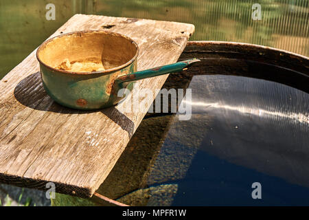 Regenfass gefüllt mit Wasser und Metallpfanne auf Holzbrett an sonnigen Tagen Stockfoto