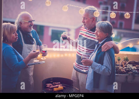 Gruppe von Freunden Senioren ein BBQ Grill zusammen auf der Dachterrasse mit herrlichem Blick auf andere Gebäude. Lampe leuchtet auf der Terrasse und Lächeln ha Stockfoto