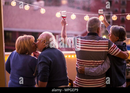 Vier 70-Jährigen auf der Terrasse Toasten mit Glas Pokale und Rotwein feiern, umarmen und küssen einander. Hängenden gelben Glühbirnen, die c Stockfoto