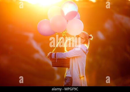 Hund und Mensch identisch ist. Schöne Mädchen trägt einen Korb mit Kugeln und Küssen eines Welpen in den Strahlen der untergehenden Sonne. Das Konzept der Freude, Glück. Stockfoto