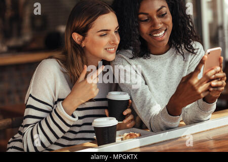 Zwei Frauen in einem Restaurant mit Blick auf Handy sitzt und spricht. Freunde sitzen in einem Café mit Kaffee und Snacks auf den Tisch unter selfie Usi Stockfoto