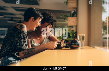 Zwei junge Frauen, die mit einem Smart Phone im Coffee Shop. Zwei lächelnde weibliche Freunde mit einem Mobiltelefon in einem Restaurant. Stockfoto