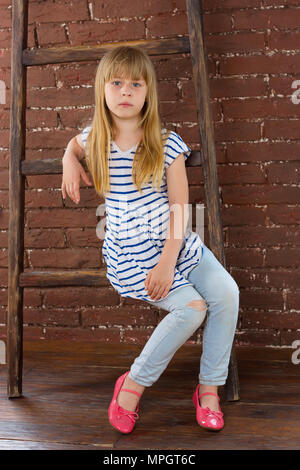 Mädchen 6 Jahre alt in Jeans sitzt auf einer Leiter in der Nähe einer Wand Stockfoto