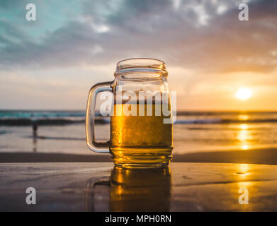 Glas Glas Bier auf einer hölzernen Tisch mit Blick auf das Meer bei Sonnenuntergang Stockfoto