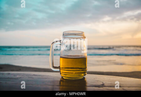 Glas Glas Bier auf einer hölzernen Tisch mit Blick auf das Meer bei Sonnenuntergang Stockfoto