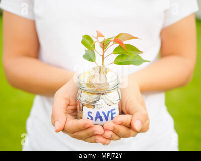 Frauen Hand, die Pflanze, die aus Münzen in Glas Glas auf dem grünen Rasen für Geld sparen Finanzkonzept Stockfoto