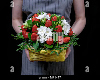 Der Florist hält einen Korb mit Blumen und Erdbeeren auf einem dunklen Hintergrund. Florist bei der Arbeit Stockfoto