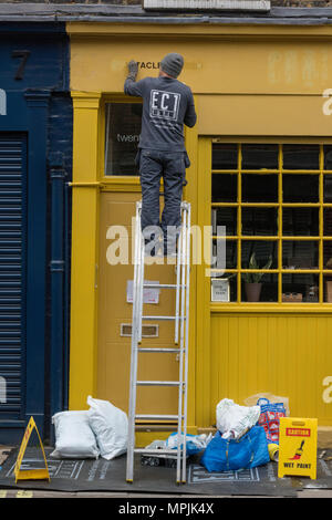 Ein Mann mit einer Leiter ein Gebäude Gelb zu malen. Mann auf eine Leiter auf der Seite eines Gebäudes Maler und Dekorateur bei der Arbeit Bild in hellen Farben. Stockfoto