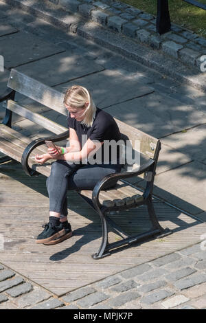 Eine junge Frau mit blondem Haar in Jeans sitzt auf einer Holzbank auf dem Bürgersteig mit einem mobilen Gerät oder Telefon, SMS oder E-Mail betrachten. Stockfoto