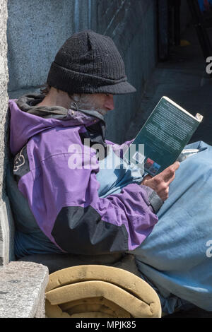 Ein obdachloser Mann sitzen auf dem Boden trägt einen schwarzen Hut und ein Buch lesen im Zentrum der Hauptstadt von London. obdachlosigkeit in der Hauptstadt. Stockfoto