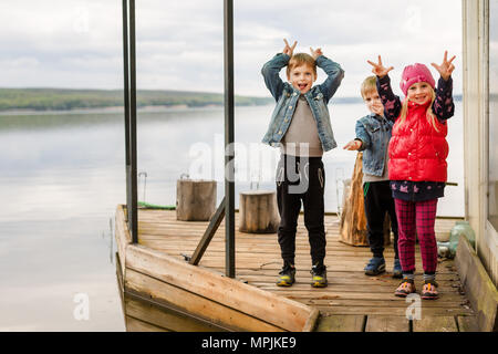 Drei Freunde spielen, angeln auf hölzernen Pier in der Nähe von Teich. Kleinkind zwei Jungen und ein Mädchen am Ufer. Kinder haben Spaß daran, Grimasse. Happy childhoo Stockfoto