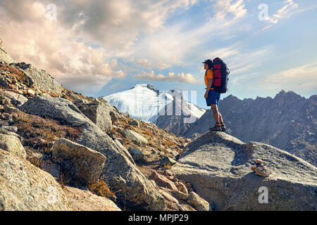 Wanderer in orangefarbenen T-Shirt mit großem Rucksack auf der Suche auf dem verschneiten Gipfel im Hintergrund Stockfoto