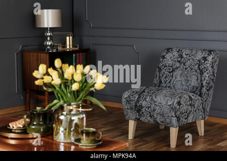 Elegantes Wohnzimmer Einrichtung mit gemusterten Sessel gegen schwarze Wand mit Spritzguss- und unscharfes gelbe Tulpen auf einem Tisch im Vordergrund. Re Stockfoto