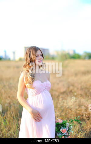 Schwangere junge Frau tragen rosa Kleid mit Blumenstrauß in Steppen Hintergrund stehen. Stockfoto