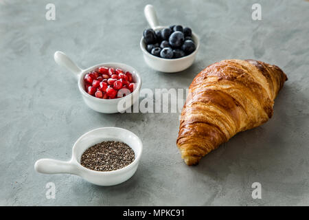 Hohen winkel Nahaufnahme einer Konditorei Anordnung - Croissants und Tassen mit Blaubeeren, Granatapfel und Chia Samen auf einem grauen Oberfläche Stockfoto