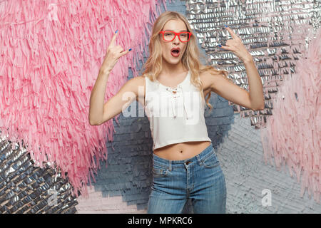 Glück junge Langhaarige blonde Mädchen in rot Gläser, Rock und Roll Finger und Kamera mit verrückt aussehen Stockfoto