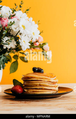 Gestapelte Pfannkuchen mit Beeren und Sirup auf Tisch mit Blumen in der Vase Stockfoto