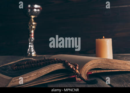 Offene Bibel mit Rosenkranz, Kelch und Kerze auf Holztisch, die Heilige Kommunion Stockfoto