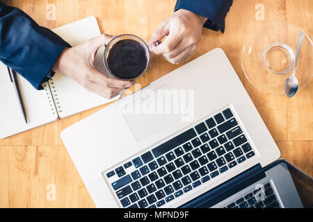 Blick von oben auf einen Geschäftsmann Hände halten eine Tasse schwarzen Kaffee in der Nähe von seinen Laptop, gut für die Arbeit leben oder Business Thema Hintergrund Stockfoto
