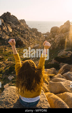 Italien, Sardinien, Frau auf einer Wanderung sitzt auf Felsen an der Küste ihre Arme anheben Stockfoto