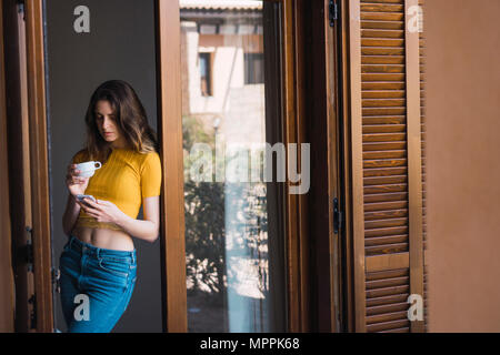 Junge Frau mit Tasse Kaffee gegen Tür Fall lehnte sich an Zelle Telefon Stockfoto