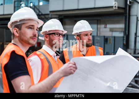 Drei Männer Tragen von Schutzhelmen und Warnwesten holding Plan in der Factory Stockfoto