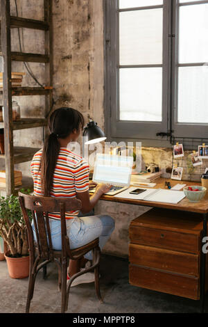 Zurück Blick auf die junge Frau am Schreibtisch sitzen in einem Loft Arbeiten am Laptop Stockfoto