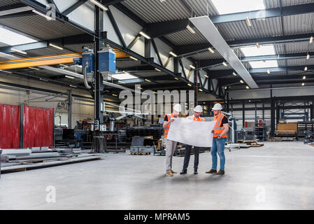 Drei Männer Tragen von Schutzhelmen und Warnwesten bei plan auf dem Shop floor suchen Stockfoto