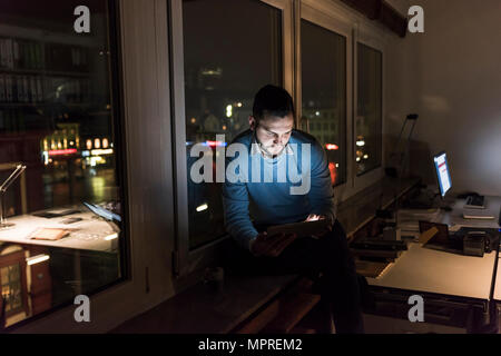 Unternehmer sitzen auf Fensterbank in Büro in der Nacht auf Tablet Stockfoto