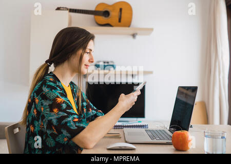 Junge Frau am Tisch zu Hause mit Handy und Laptop Stockfoto