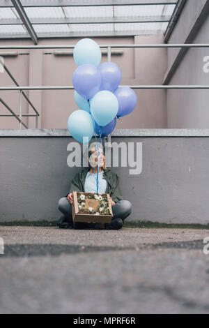 Lächelnde Frau mit Blumen im Karton und Bündel von blauen Ballons sitzen auf dem Boden Stockfoto