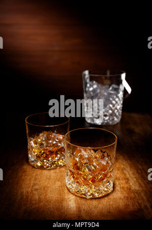 Ein Schlaglicht auf zwei Kristall Glas Scotch Whisky mit einem Eiskübel im backghround, Schuß auf einen hölzernen Tisch. Stockfoto