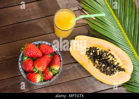 In der Hälfte reife Papaya frische Erdbeeren tropischen Saft in hohes Glas mit Stroh. Palm Leaf auf der Plank Holz Tisch im Café. Urlaub Sommer Reisen Con Stockfoto